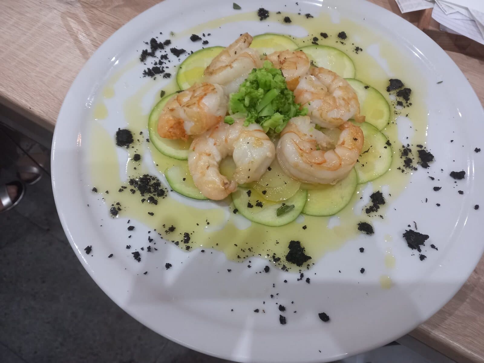Garnelen auf Zucchini-Carpaccio mit Olivenöl, Erbsencreme und dunklem Krokant, angerichtet auf weissem Teller